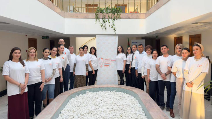 A group of people wearing white shirts stand in a semicircle around a stone-filled fountain, posing indoors beside a banner about activism against gender-based violence.